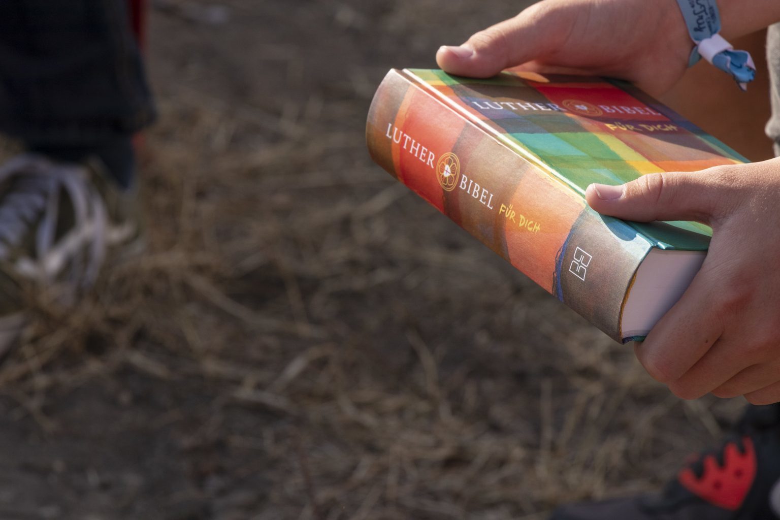 Eine Person, die eine bunte, gebundene Ausgabe der Lutherbibel in der Hand hält, mit trockenem Gras im Hintergrund und den Schuhen einer anderen Person, die teilweise sichtbar ist - ein lebendiger Moment, der bei den evangelischen Jugendbremen (ejhb) in Bremen aufgenommen wurde.
