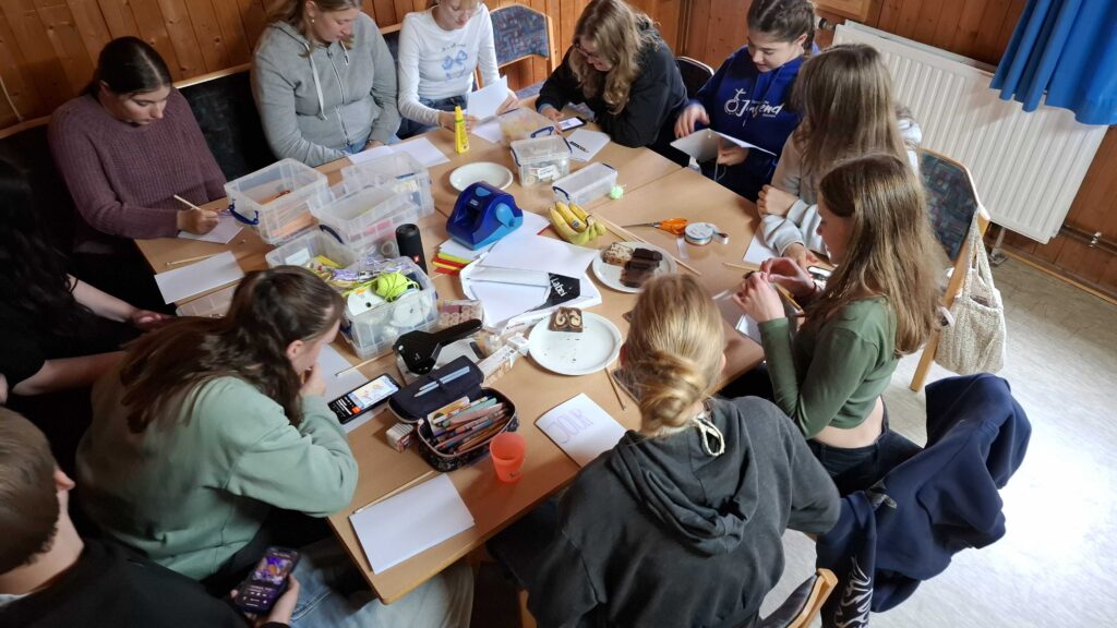 Eine Gruppe von Menschen sitzt in einem gemütlichen, holzgetäfelten Raum um einen Tisch herum, auf dem Papier, Stifte, Marker und anderes Material verstreut sind, und beschäftigt sich mit Kunsthandwerk.