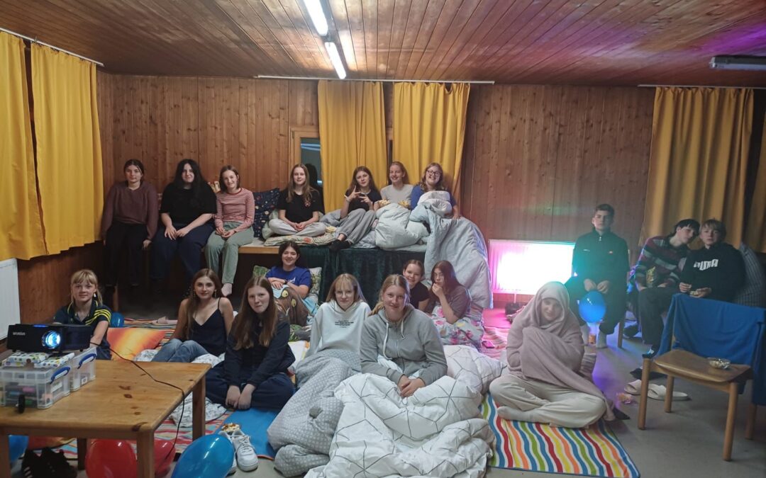 Eine Gruppe junger Leute sitzt eng beieinander auf Decken und Matratzen in einem hölzernen Raum mit gelben Vorhängen, bunten Luftballons und einem Projektor, der an eine entspannte Film- oder Übernachtungsnacht erinnert.