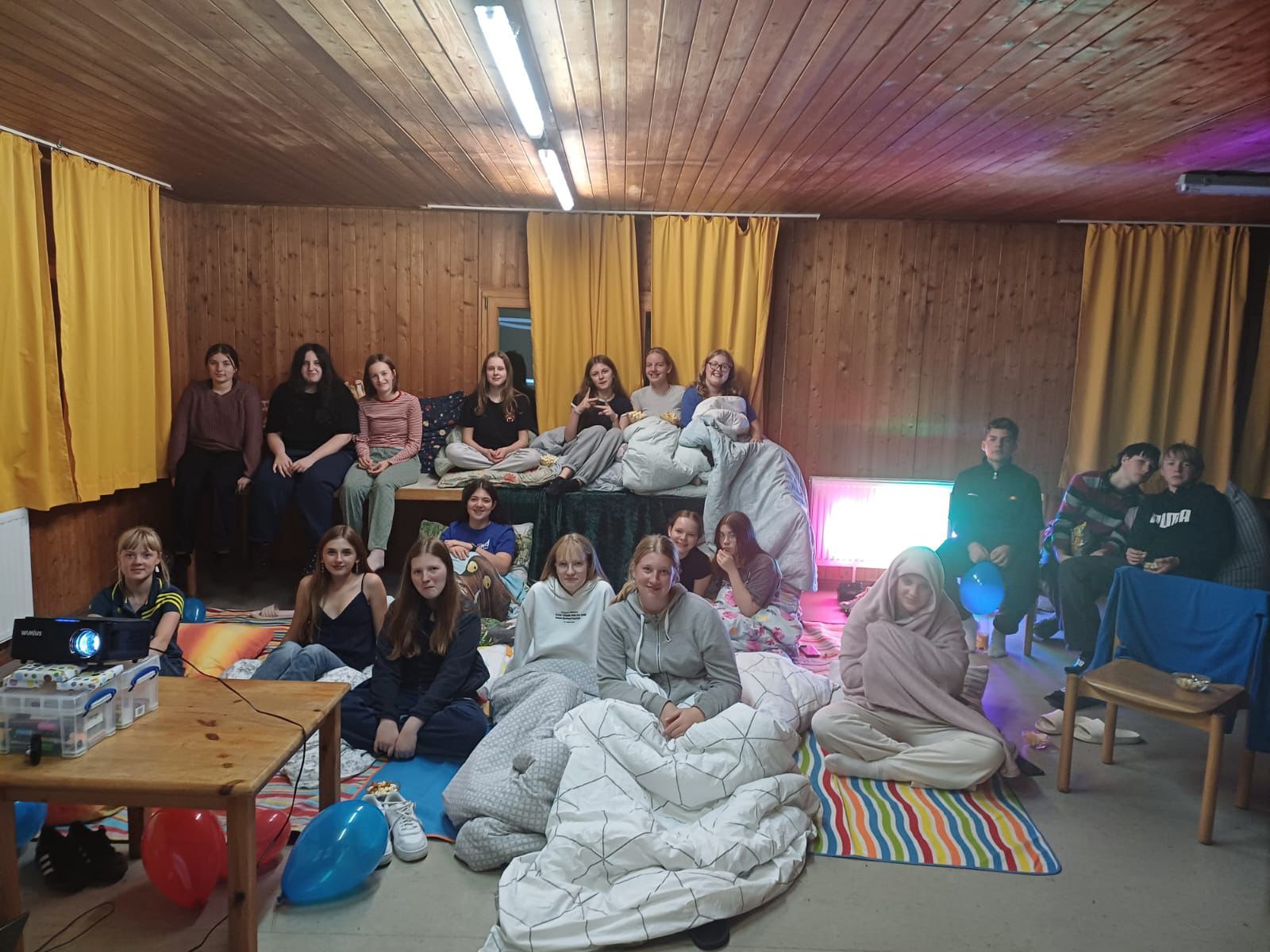 Eine Gruppe junger Leute sitzt eng beieinander auf Decken und Matratzen in einem hölzernen Raum mit gelben Vorhängen, bunten Luftballons und einem Projektor, der an eine entspannte Film- oder Übernachtungsnacht erinnert. Eine Gruppe junger Leute sitzt eng beieinander auf Decken und Matratzen in einem hölzernen Raum mit gelben Vorhängen, bunten Luftballons und einem Projektor, der an eine entspannte Film- oder Übernachtungsnacht erinnert.