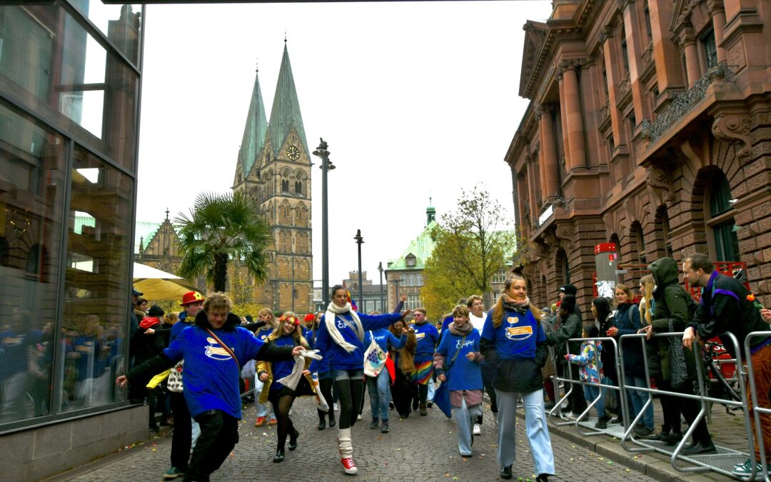 Eine Gruppe von Menschen in blauen Hemden geht lächelnd eine von jubelnden Schaulustigen gesäumte Straße entlang; im Hintergrund sind unter einem bedeckten Himmel historische Gebäude und eine hohe Kathedrale zu sehen.