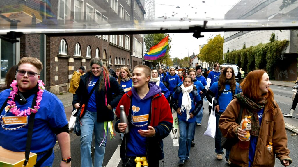 Eine Gruppe von Menschen in blauen Hemden und mit bunten Accessoires marschiert eine Straße entlang, einige halten eine Regenbogenfahne in der Hand. Sie wirken fröhlich und energiegeladen und nehmen an einer lebhaften öffentlichen Veranstaltung oder Parade teil.