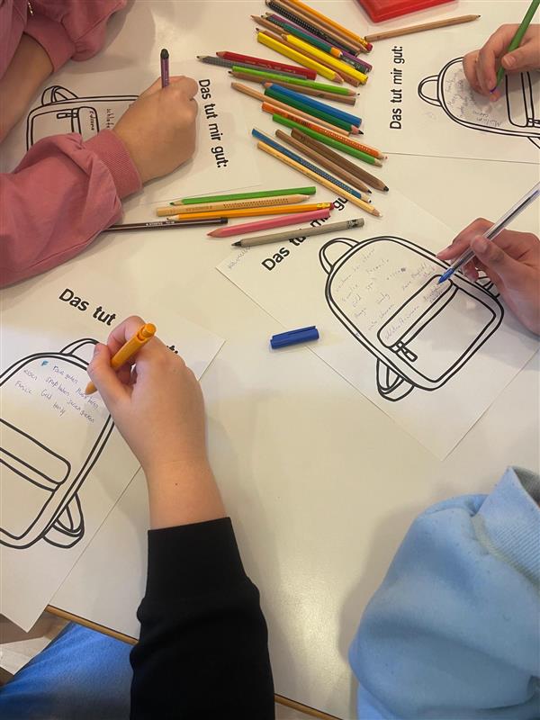Vier Kinder sitzen um einen Tisch und malen und schreiben auf Arbeitsblättern mit einer Rucksackillustration und dem deutschen Satz Das tut mir gut. Buntstifte liegen verstreut auf dem Tisch.