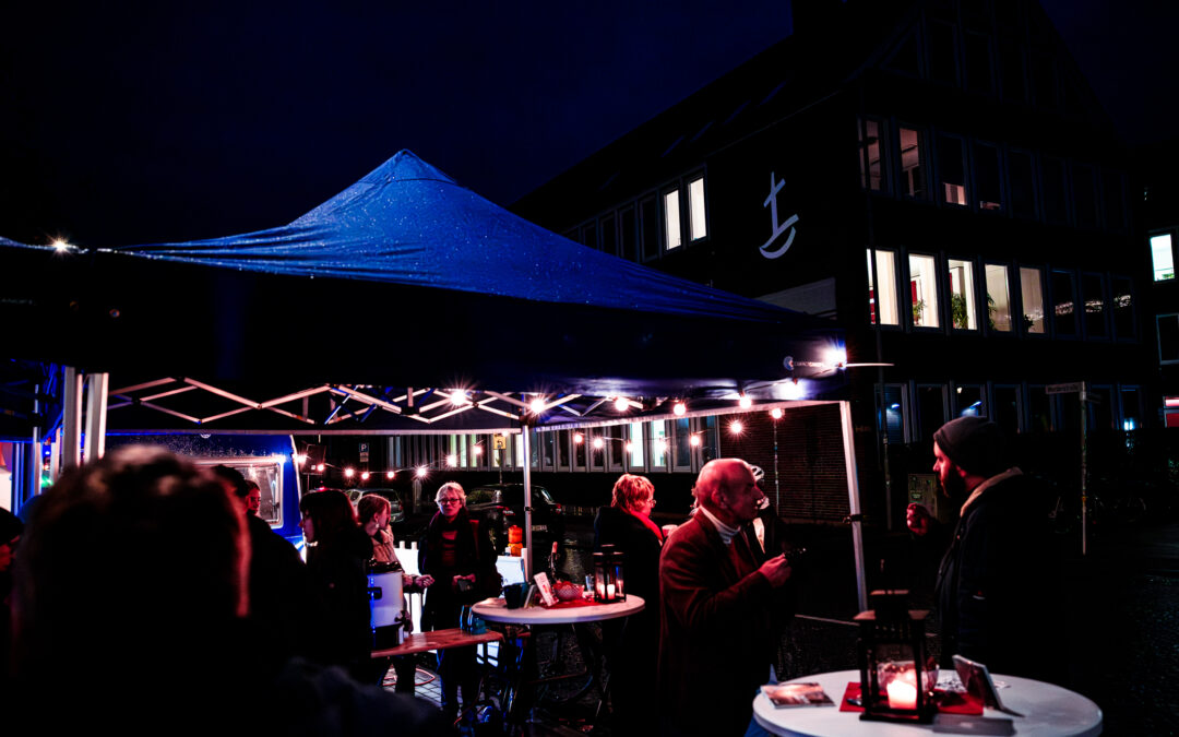 Menschen versammeln sich nachts an runden Tischen unter einem blauen Vordach mit Lichterketten, unterhalten sich und trinken. Im Hintergrund ist ein dunkles Gebäude mit beleuchteten Fenstern und einem weißen Ankersymbol an der Fassade zu sehen.