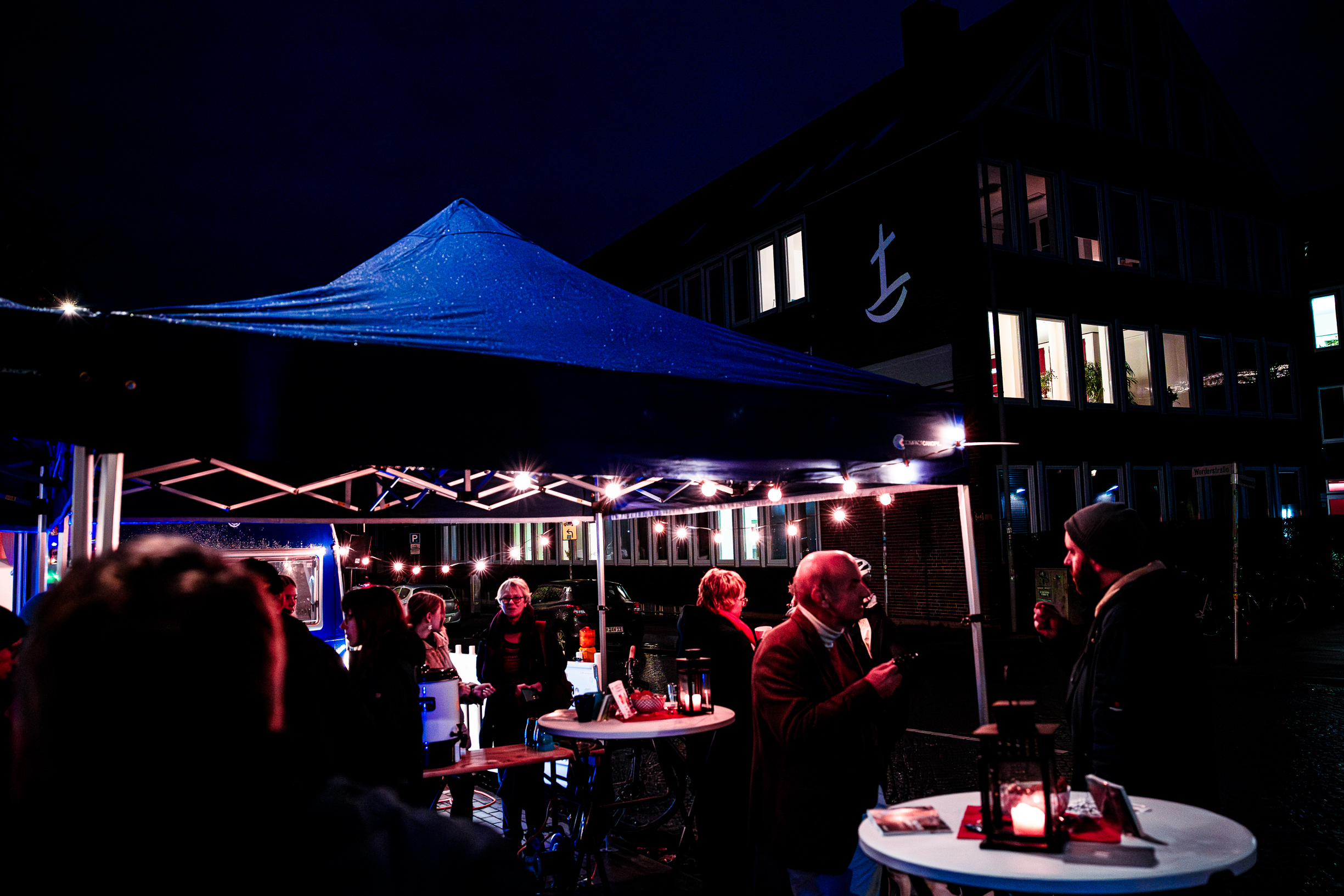 Menschen versammeln sich nachts an runden Tischen unter einem blauen Vordach mit Lichterketten, unterhalten sich und trinken. Im Hintergrund ist ein dunkles Gebäude mit beleuchteten Fenstern und einem weißen Ankersymbol an der Fassade zu sehen. Menschen versammeln sich nachts an runden Tischen unter einem blauen Vordach mit Lichterketten, unterhalten sich und trinken. Im Hintergrund ist ein dunkles Gebäude mit beleuchteten Fenstern und einem weißen Ankersymbol an der Fassade zu sehen.