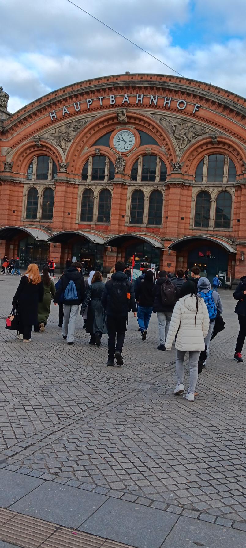 Eine Gruppe von Menschen geht an einem bewölkten Tag auf den historischen Hauptbahnhof aus rotem Backstein mit Bogenfenstern, einer großen Uhr und dem Schild HAUPTBAHNHOF über dem Eingang zu. Eine Gruppe von Menschen geht an einem bewölkten Tag auf den historischen Hauptbahnhof aus rotem Backstein mit Bogenfenstern, einer großen Uhr und dem Schild HAUPTBAHNHOF über dem Eingang zu.