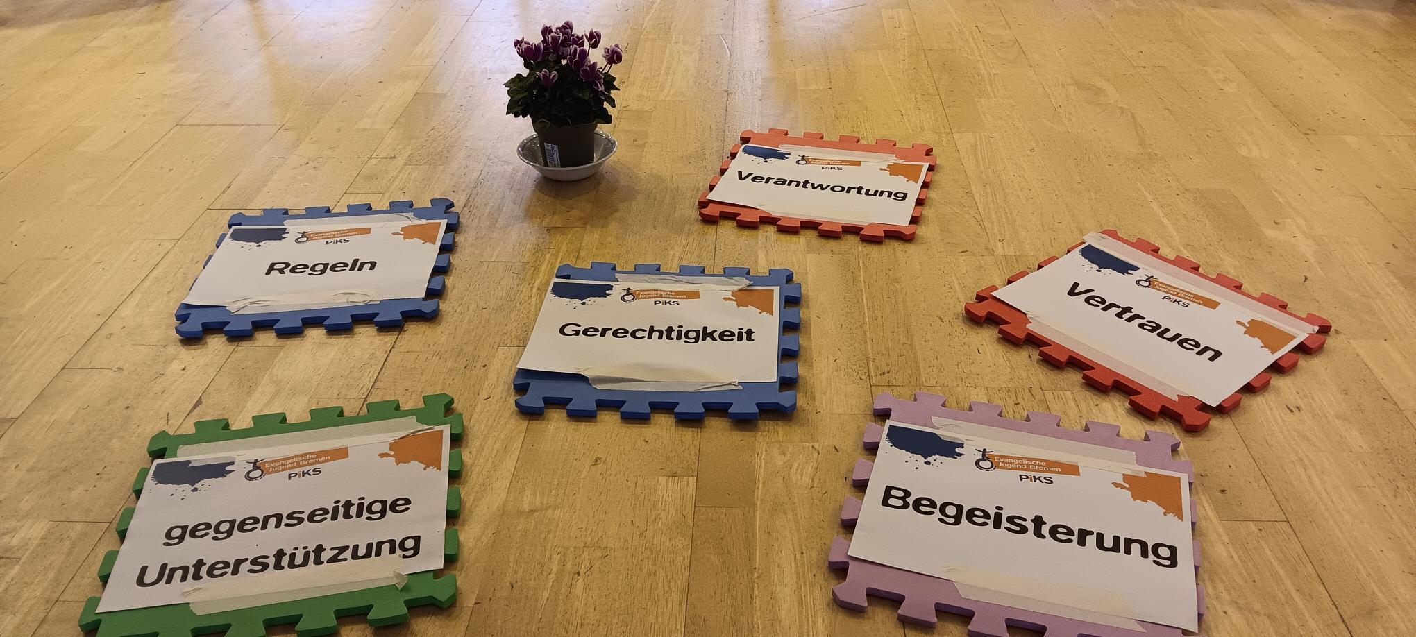 Sechs bunte Schaumstoffmatten mit deutschen Wörtern (Regeln, gegenseitige Unterstützung, Gerechtigkeit, Verantwortung, Vertrauen, Begeisterung) sind im Kreis auf einem Holzboden angeordnet, in der Mitte steht eine Topfpflanze.