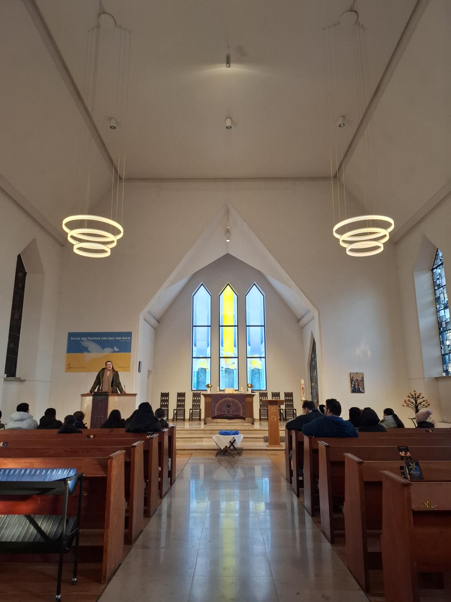 Ein modernes Kircheninnere mit hohen Decken, Holzbänken und mit Blick auf den Altar sitzenden Menschen. Eine Person steht an der Kanzel, und große Buntglasfenster in Blau und Gelb erhellen den Altarbereich.