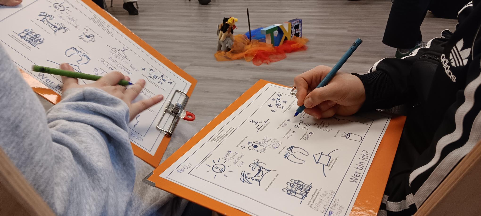 Zwei Personen, die Klemmbretter in der Hand halten, zeichnen Bilder auf Arbeitsblättern. Auf den Arbeitsblättern sind handgezeichnete Kritzeleien und Wörter zu sehen. Im Hintergrund sind kleine Figuren, bunte Stoffe und Schachteln auf dem Boden zu sehen.