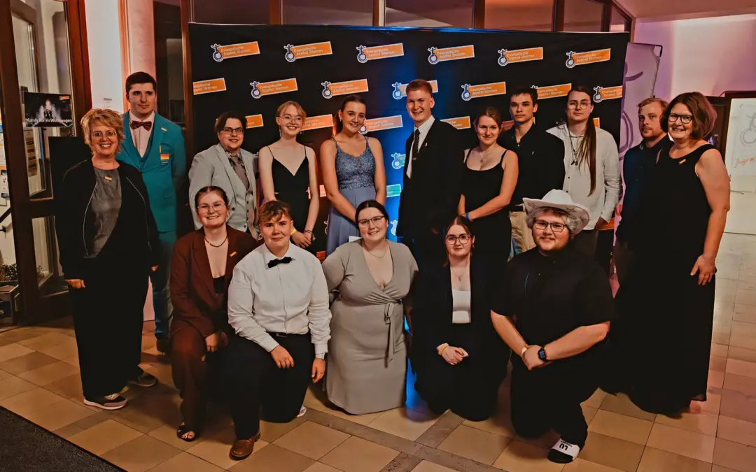 Eine Gruppe formell und semi-formell gekleideter Personen posiert lächelnd vor einem schwarzen Step-and-Repeat-Banner bei einer Indoor-Veranstaltung mit warmer Beleuchtung.