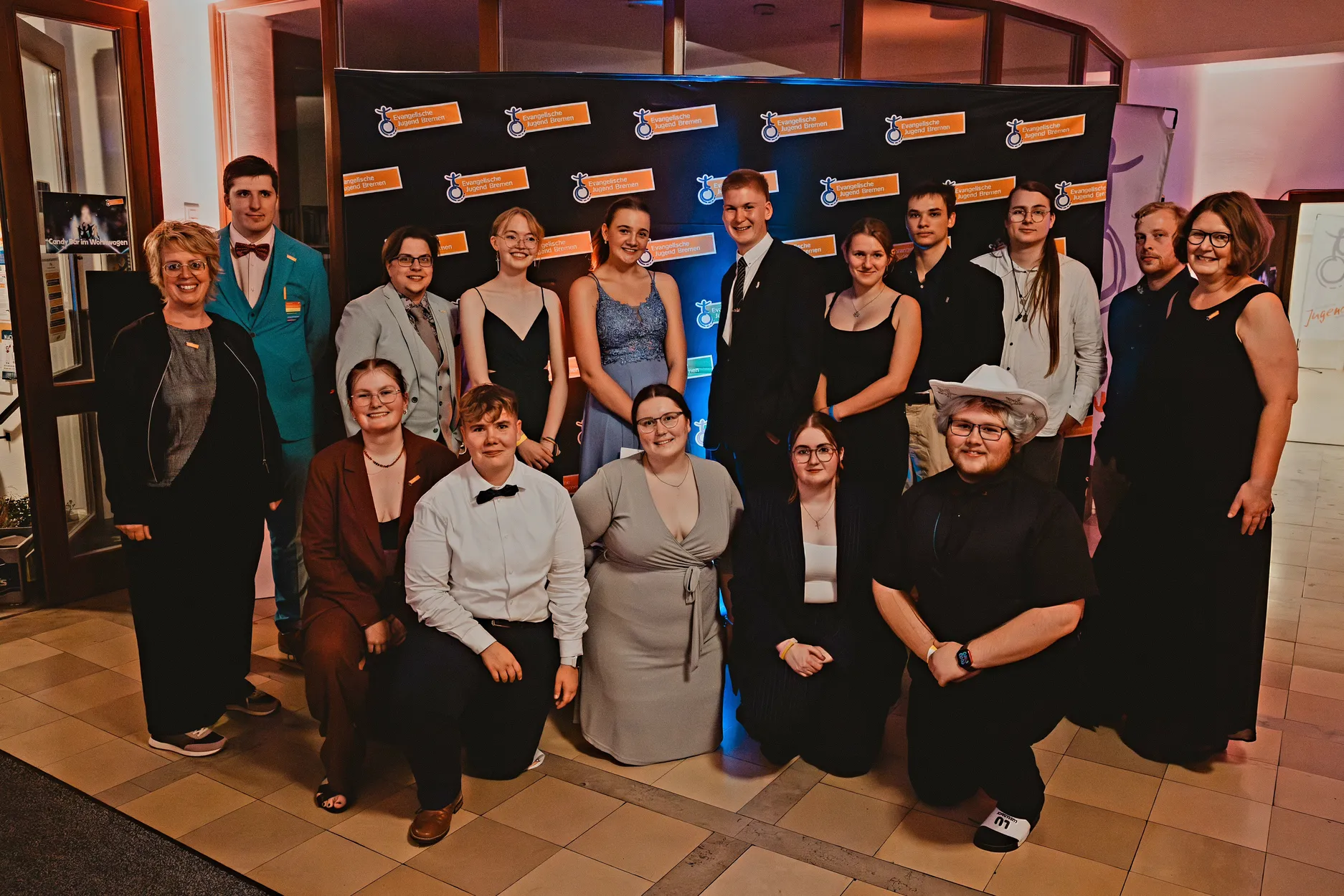 Eine Gruppe formell und semi-formell gekleideter Personen posiert lächelnd vor einem schwarzen Step-and-Repeat-Banner bei einer Indoor-Veranstaltung mit warmer Beleuchtung.