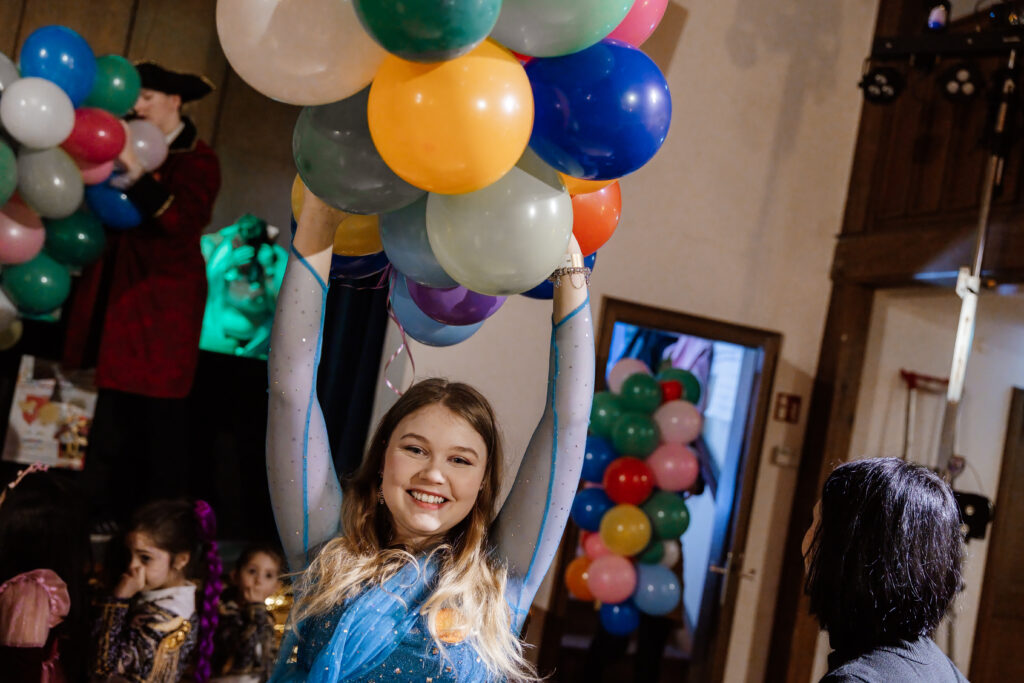 Eine lächelnde junge Frau in einem blauen Kleid hält bunte Luftballons über ihrem Kopf auf einer Indoor-Party, während im Hintergrund andere Menschen und weitere Ballons zu sehen sind.