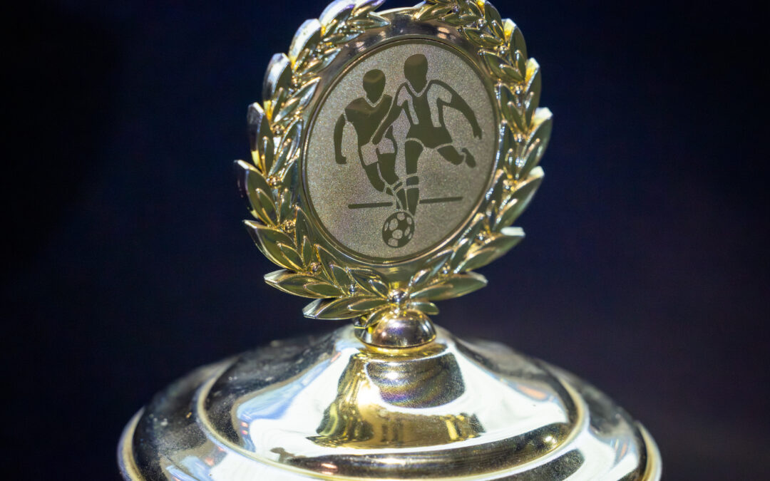 Nahaufnahme eines glänzenden goldenen Fußballpokals mit einem runden Emblem, das zwei Fußballspieler und einen Ball zeigt, eingerahmt von einem Lorbeerkranz, vor einem dunklen Hintergrund.