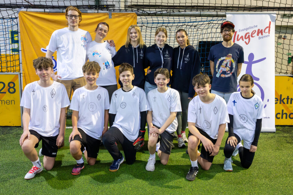 Eine Gruppe von acht Jugendlichen in weißen Sportuniformen kniet oder steht vorne, fünf Erwachsene in Freizeitkleidung stehen hinter ihnen auf dem Hallenrasen. Im Hintergrund sind Fahnen und Netze zu sehen. Alle lächeln in die Kamera.