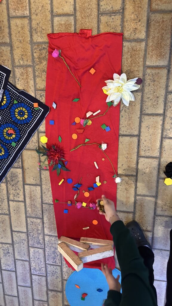 Eine Kinderhand greift nach einem roten Tuch auf einem Backsteinboden, der mit Blumen, bunten Holzformen und Holzklötzen verziert ist. In der Nähe liegen ein gemusterter Stoff und ein blauer Kreis.