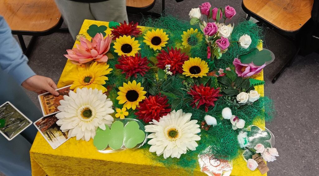 Ein gelbes Tischtuch bedeckt einen mit Kunstblumen in Rot, Weiß, Gelb und Rosa geschmückten Tisch, der von grünem Kunstgras umgeben ist. Auf dem Tisch liegen auch ein paar Kleeblätter und Postkarten aus Papier.