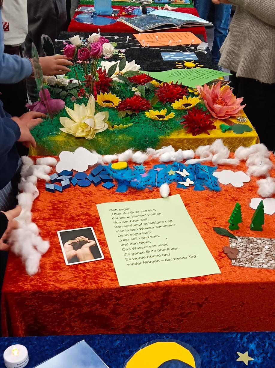 Ein farbenfrohes Display auf einem Tisch zeigt künstliche Blumen, Baumwollwolken, Bäume und Wasser, dazu ein grünes Papier mit deutschem Text und ein Foto von zum Gebet gefalteten Händen. Mehrere Menschen stehen in der Nähe.