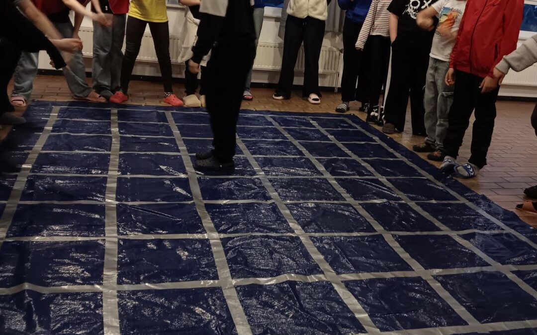 Eine Gruppe von Personen steht in einem Kreis um eine große blaue Plane auf dem Boden, die mit einem Gitter aus Klebeband markiert ist; eine Person steht in der Mitte des Gitters, während die anderen zusehen.