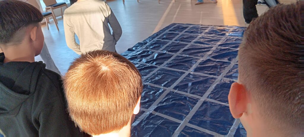 Drei Kinder stehen in einem Haus und blicken auf eine große blaue Plane auf dem Boden, die mit einem Gitter aus weißem Klebeband markiert ist. Das Sonnenlicht fällt von der Seite ein und wirft Schatten auf die Szene.