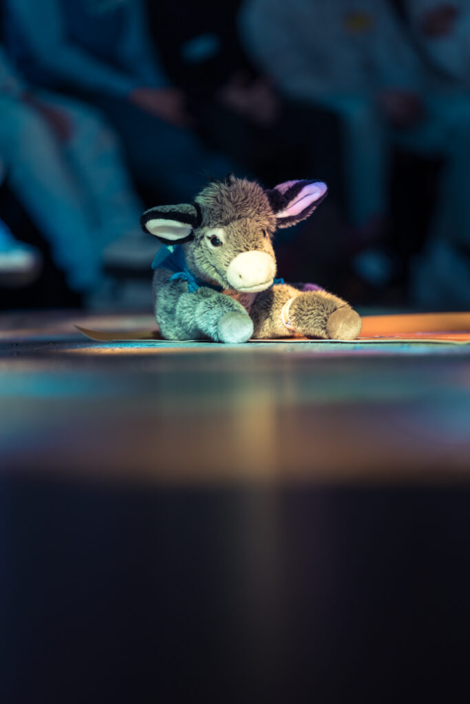 Ein Plüschesel sitzt auf einer Bühne, die von einer sanften, bunten Beleuchtung hervorgehoben wird, mit unscharfen Menschen im Hintergrund.