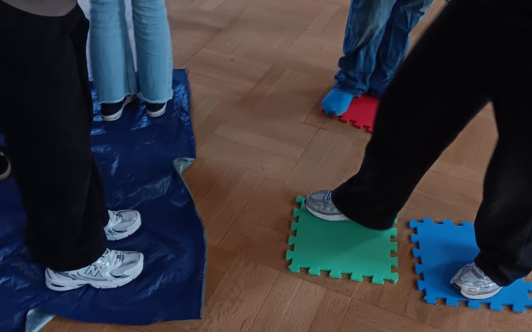 Mehrere Personen stehen auf Schaumstoffmatten und einer blauen Plane, die auf einem Holzboden liegt. Nur ihre Beine und Füße sind zu sehen. Sie tragen alle Freizeitkleidung und Turnschuhe, eine Person hat blaue Socken an.