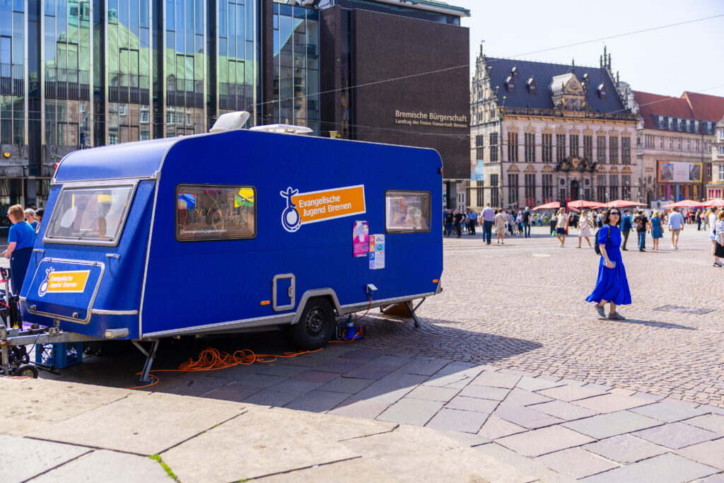 Ein blauer Wohnwagen mit Schildern der Evangelischen Jugend Bremen steht an einem sonnigen Tag auf einem belebten Europaplatz mit Spaziergängern und historischen Gebäuden im Hintergrund.