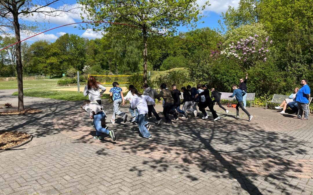An einem sonnigen Tag rennt eine Gruppe von Kindern gemeinsam in einem Park, im Hintergrund sind grüne Bäume und ein blauer Himmel zu sehen. Einige Erwachsene beobachten sie von nahegelegenen Bänken aus. Die Schatten der Äste fallen über den Weg.