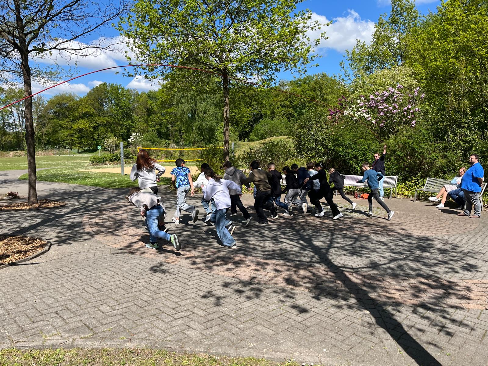 An einem sonnigen Tag rennt eine Gruppe von Kindern gemeinsam in einem Park, im Hintergrund sind grüne Bäume und ein blauer Himmel zu sehen. Einige Erwachsene beobachten sie von nahegelegenen Bänken aus. Die Schatten der Äste fallen über den Weg.