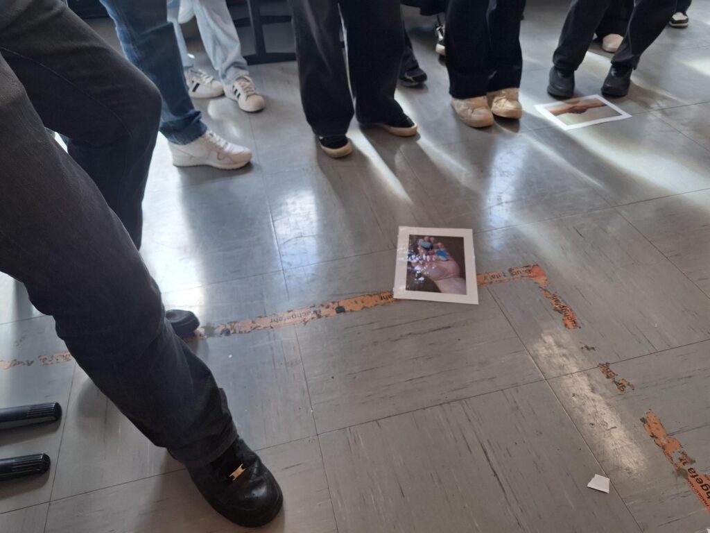 Eine Gruppe von Menschen steht in einem Raum, wobei nur ihre Beine und Schuhe zu sehen sind. Auf dem Boden liegen Fotos, und einige Personen stehen in der Nähe der Fotos oder stehen direkt darauf. Über den Boden verläuft eine abgenutzte orangefarbene Linie.