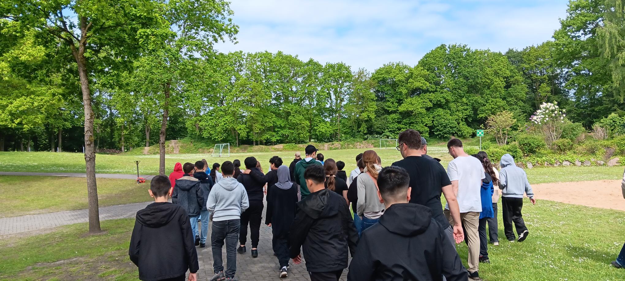 Eine Gruppe von Menschen, hauptsächlich Kinder, spaziert gemeinsam auf einem gepflasterten Weg in einem Park, der von grünen Bäumen und Gras umgeben ist, unter einem teilweise bewölkten Himmel.
