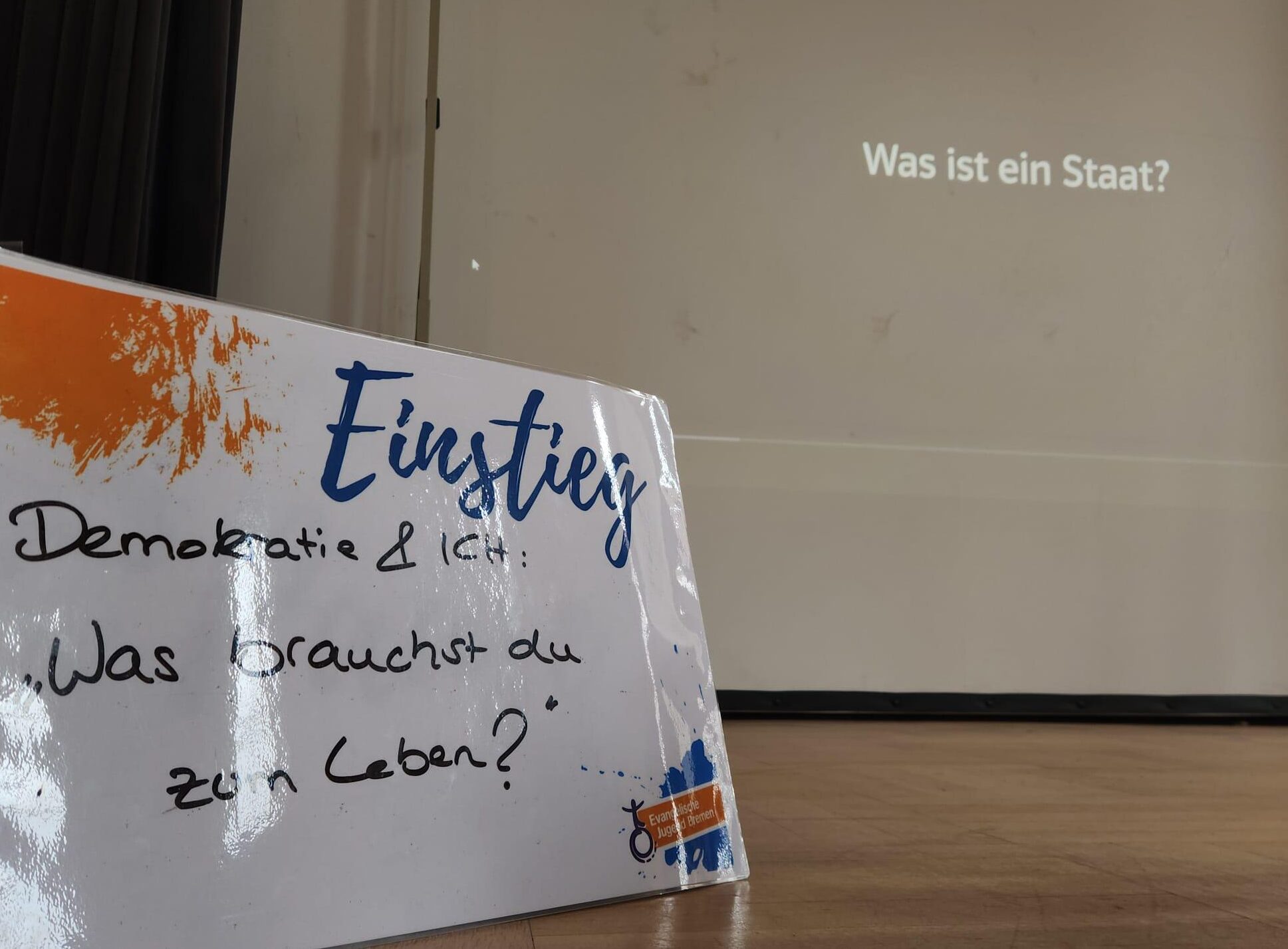 Auf dem Bühnenboden steht auf einem Schild: „Einstieg: Demokratie & Ich: Was brauchst du zum Leben?“ Im Hintergrund wird auf einer Leinwand die Frage „Was ist ein Staat?“ angezeigt. Auf dem Bühnenboden steht auf einem Schild: „Einstieg: Demokratie & Ich: Was brauchst du zum Leben?“ Im Hintergrund wird auf einer Leinwand die Frage „Was ist ein Staat?“ angezeigt.