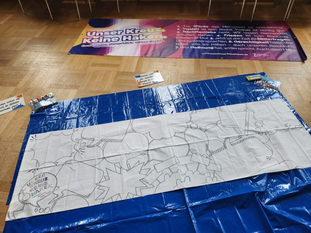 Auf einer blauen Plane liegt eine Leinwand mit einem konturierten abstrakten Motiv, umgeben von bunten Filzstiften. Dahinter hängt ein Banner mit der deutschen Aufschrift „Unser Kreuz – Keine Haken“, daneben liegen auf einem Holzboden Informationsschilder und Flugblätter.