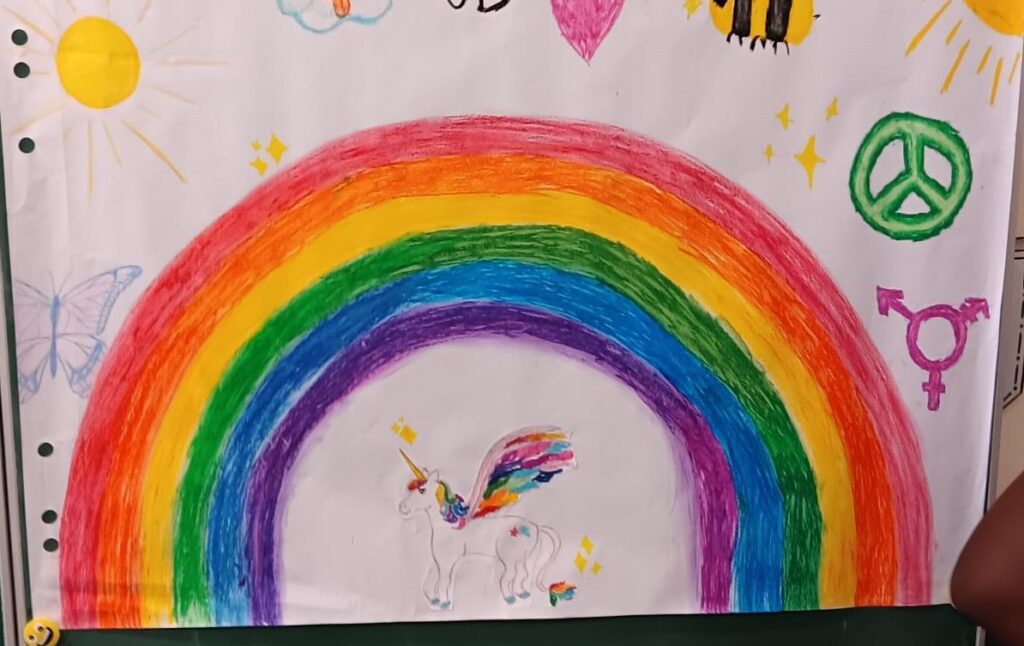 Eine farbenfrohe Zeichnung mit einem großen Regenbogen, einem Einhorn mit Regenbogenflügeln und -schwanz, einem Peace-Zeichen, einem Transgender-Symbol, einer Sonne, Wolken, einer Biene, einem Schmetterling und funkelnden Akzenten.