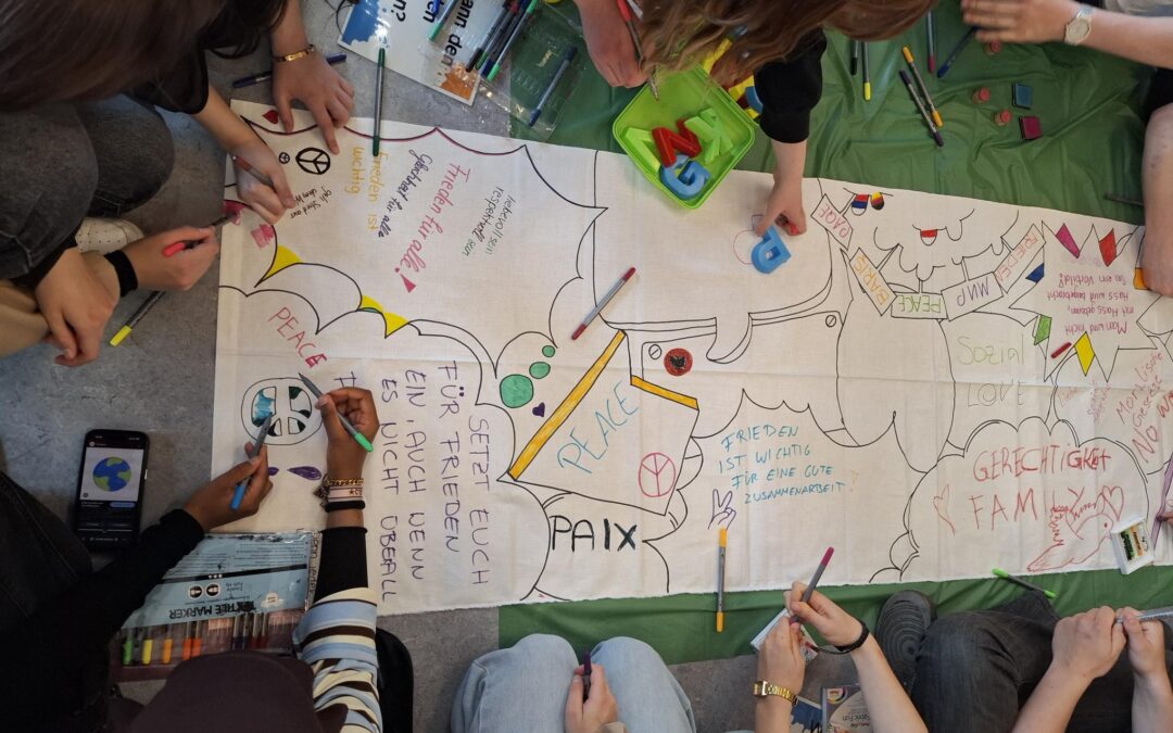Eine Gruppe von Menschen sitzt im Kreis auf dem Boden, malt und schreibt Wörter wie „peace“ und „paix“ auf ein großes Blatt Papier, während um sie herum Zeichnungen, Symbole und bunte Filzstifte verstreut liegen.