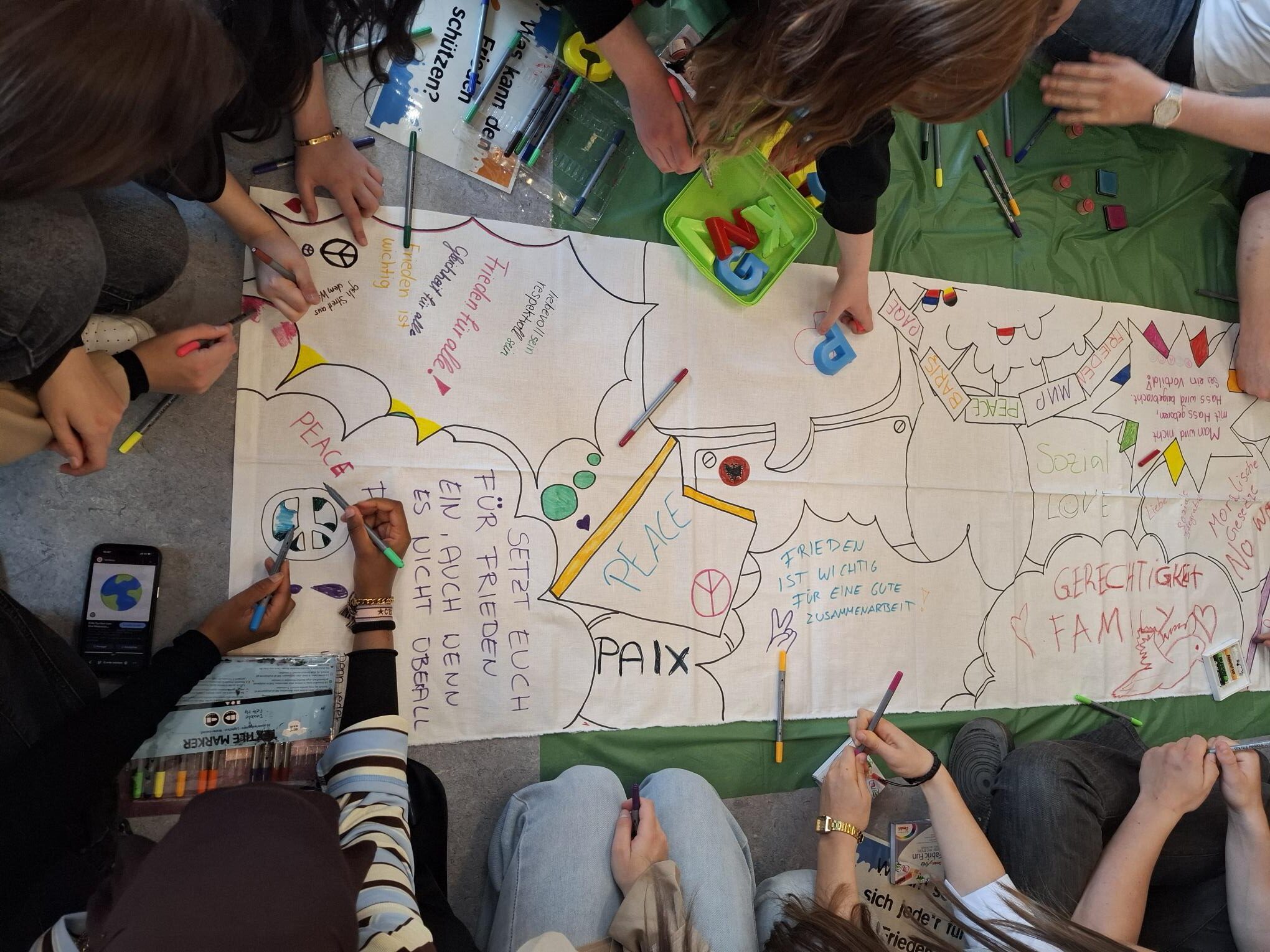 Eine Gruppe von Menschen sitzt im Kreis auf dem Boden, malt und schreibt Wörter wie „peace“ und „paix“ auf ein großes Blatt Papier, während um sie herum Zeichnungen, Symbole und bunte Filzstifte verstreut liegen.