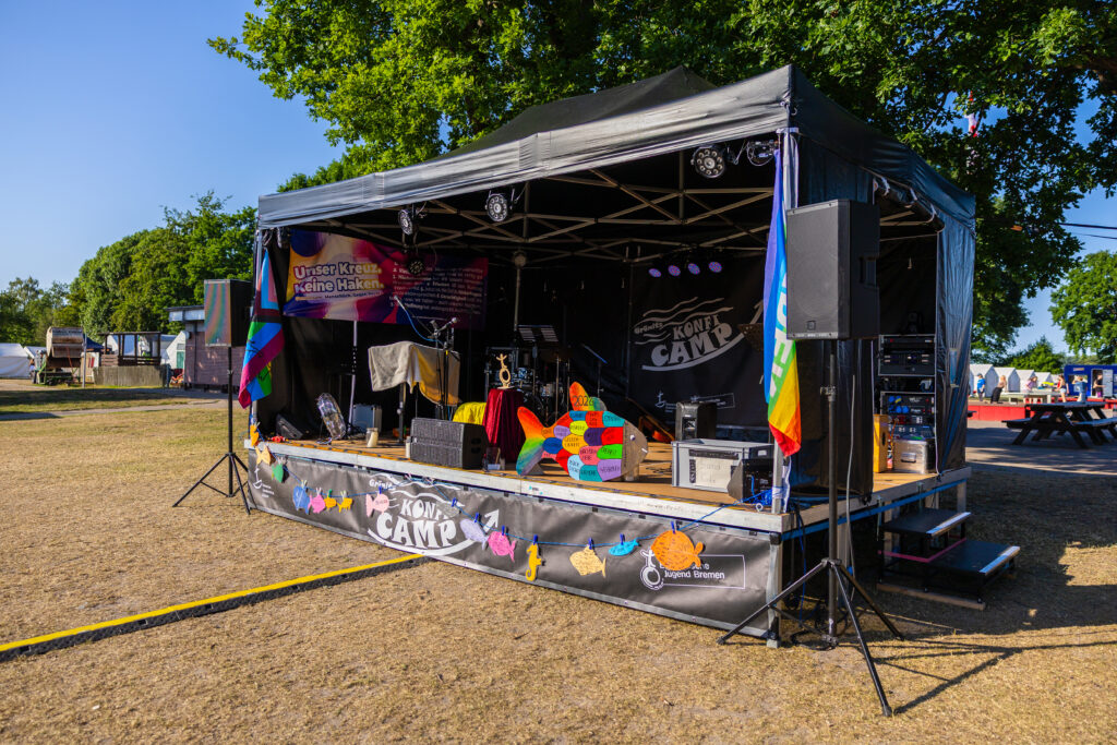 An einem sonnigen Tag steht unter einem Baum eine kleine Freiluftbühne mit Musikausrüstung, bunter Dekoration und einem KCMY CAMP-Banner. Schilder und Regenbogenfahnen sind zu sehen, was auf ein festliches Camp oder eine Gemeinschaftsveranstaltung hindeutet.
